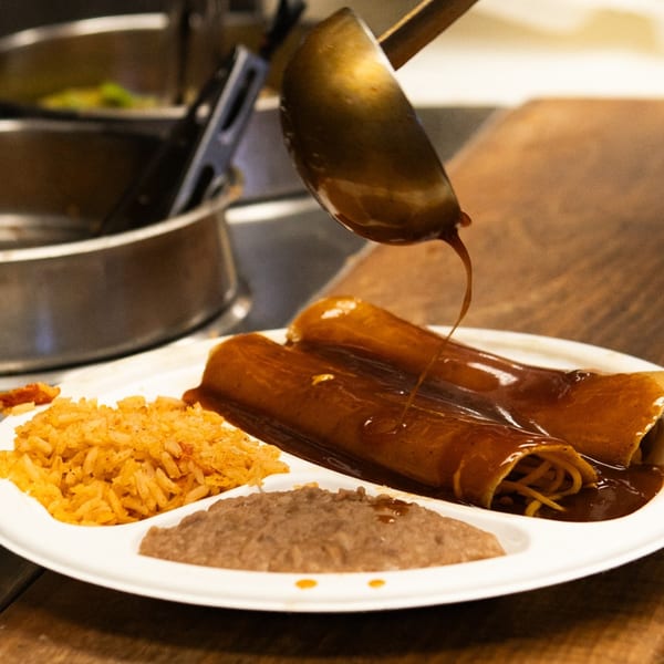 A ladle pours rich brown sauce over two enchiladas, rice, and refried beans on a white plate.